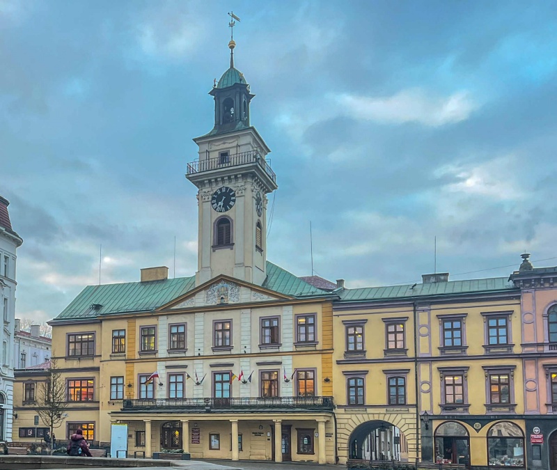 Rynek z ratuszem. Jasny budynek z wieżą i zegarem, na dole podcienia. Przed nim pusty plac.